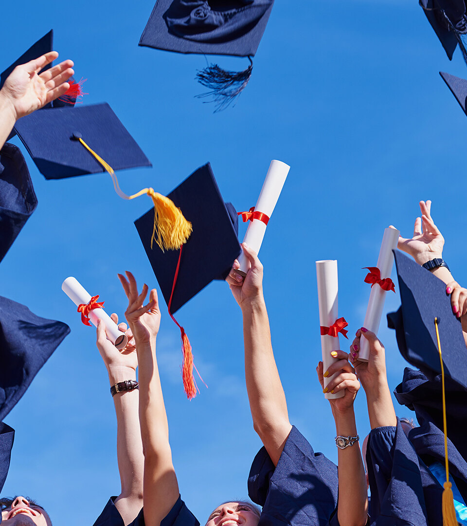 Graduating students in blue gowns celebrating with diplomas and caps under a clear blue sky