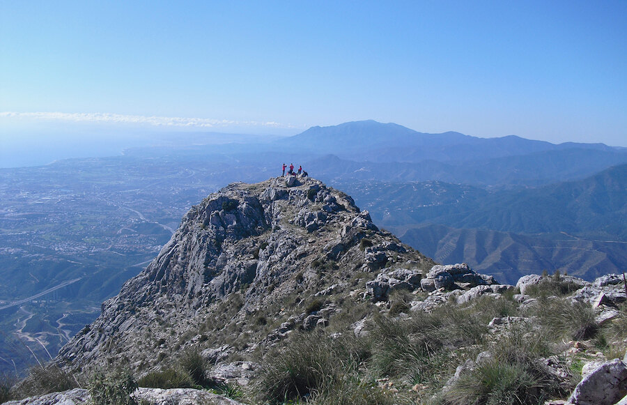 Hikers standing on a rugged mountain summit with panoramic views of the mountains and Mediterranean coastline.