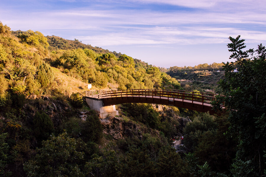 Wooden footbridge crossing a green mountain ravine surrounded by Mediterranean vegetation.