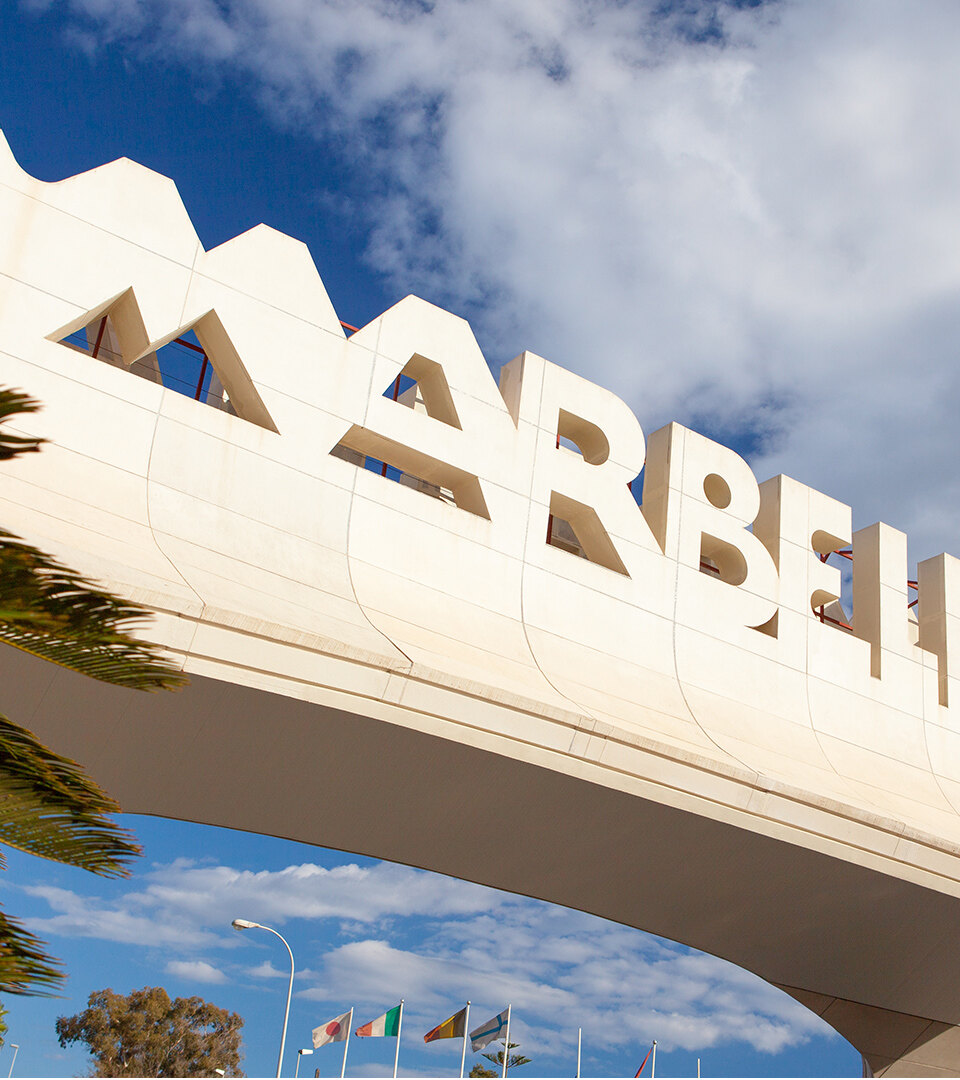 Marbella entrance arch framed by palm trees under a partly cloudy sky