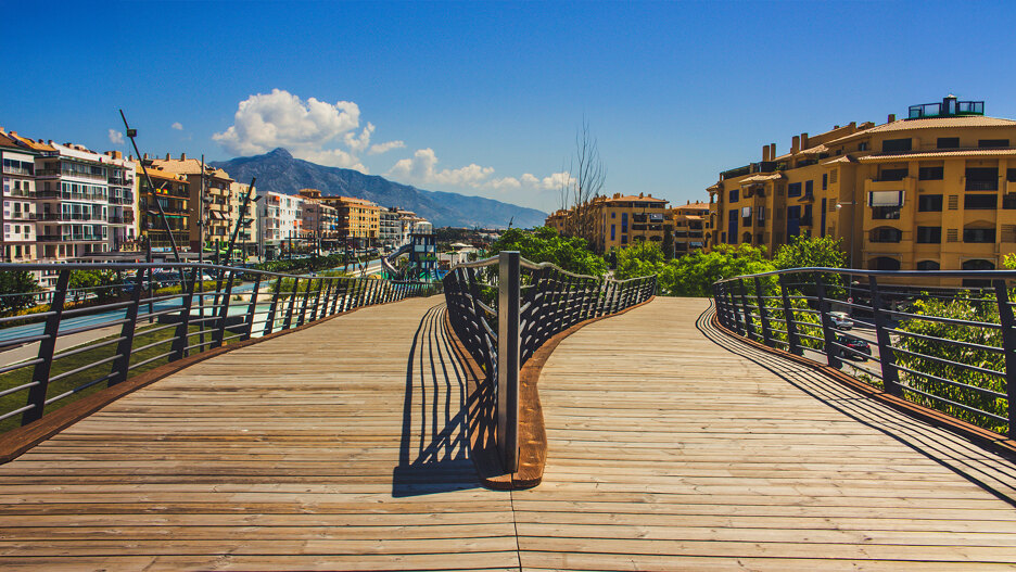 Moderne Fußgängerbrücke über den Boulevard von San Pedro de Alcántara mit dem Berg La Concha im Hintergrund, die den Kontrast zwischen Stadterneuerung und der Geschichte von San Pedro de Alcántara widerspiegelt.