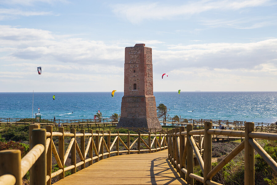 Historic watchtower by the sea in Marbella, with a wooden boardwalk and kitesurfers over the Mediterranean