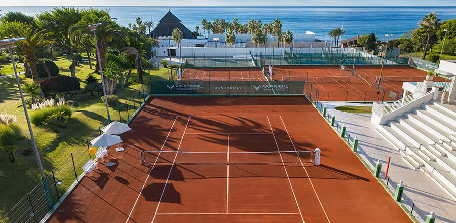 Courts en terre battue du Rafa Nadal Tennis Center Marbella, entourés de jardins de palmiers avec la mer Méditerranée en arrière-plan.