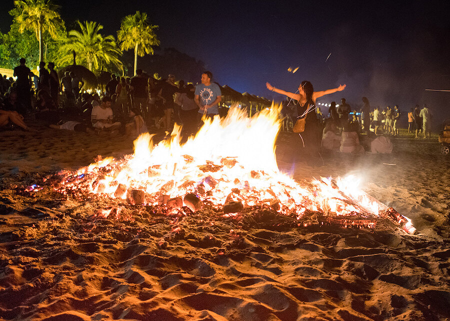 Large bonfire on the sand surrounded by people and palm trees during San Juan night in Marbella.