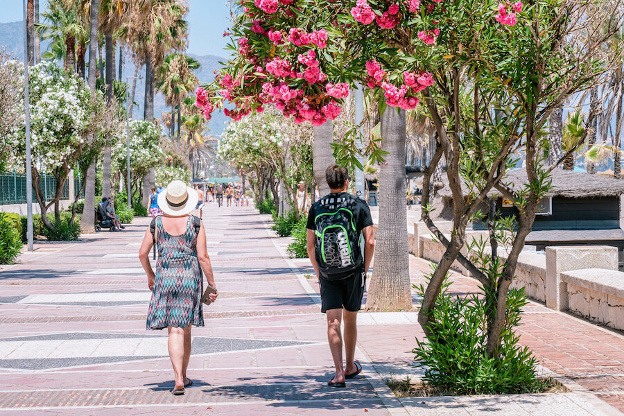 Personas caminando por un soleado paseo marítimo junto a la playa, destacando cómo las ciudades para moverse a pie crean agradables recorridos peatonales junto al mar