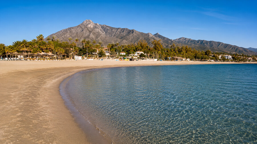 Panoramic view of a curved beach with crystal-clear water and mountain scenery in Marbella.