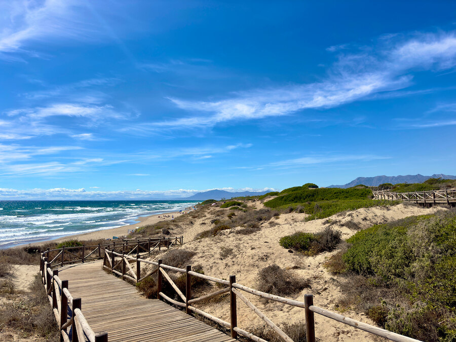 Wooden boardwalk through sandy dunes leading to a natural beach on the Costa del Sol under a bright blue sky.
