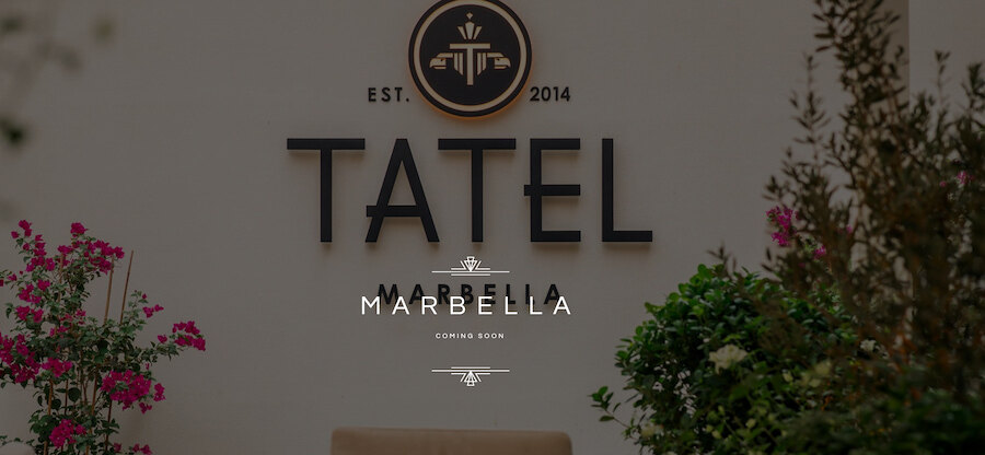 Buitenbord aan de muur van TATEL Marbella met het logo van het restaurant en de tekst “binnenkort geopend”, omlijst door groen en roze bloemen.