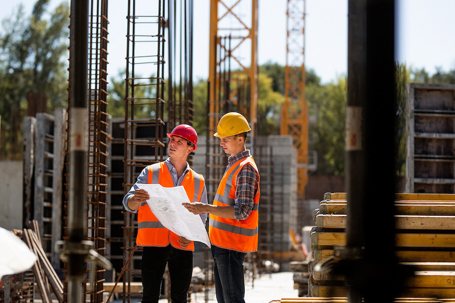 Two construction professionals in safety helmets and orange high-visibility vests reviewing plans at an active building site.