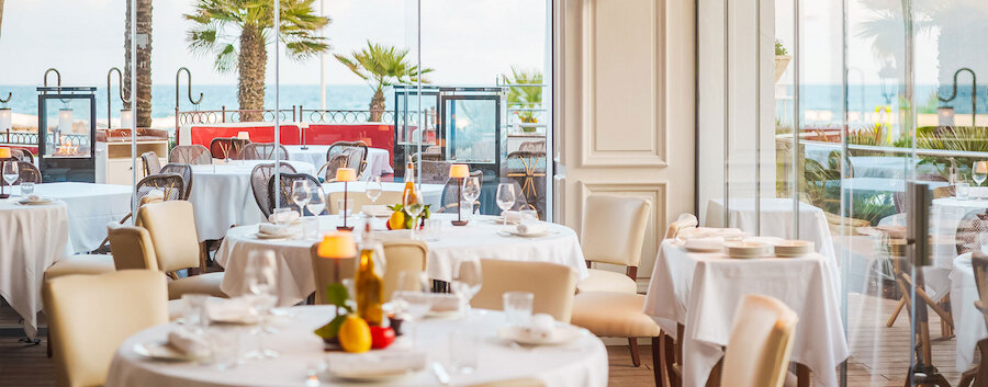 Nieuw in Marbella. Elegant restaurantinterieur aan het strand met ronde tafels met witte tafelkleden, comfortabele zitplaatsen in neutrale tinten, tafeldekking bij kaarslicht en uitzicht op zee door grote ramen, met palmbomen buiten.
