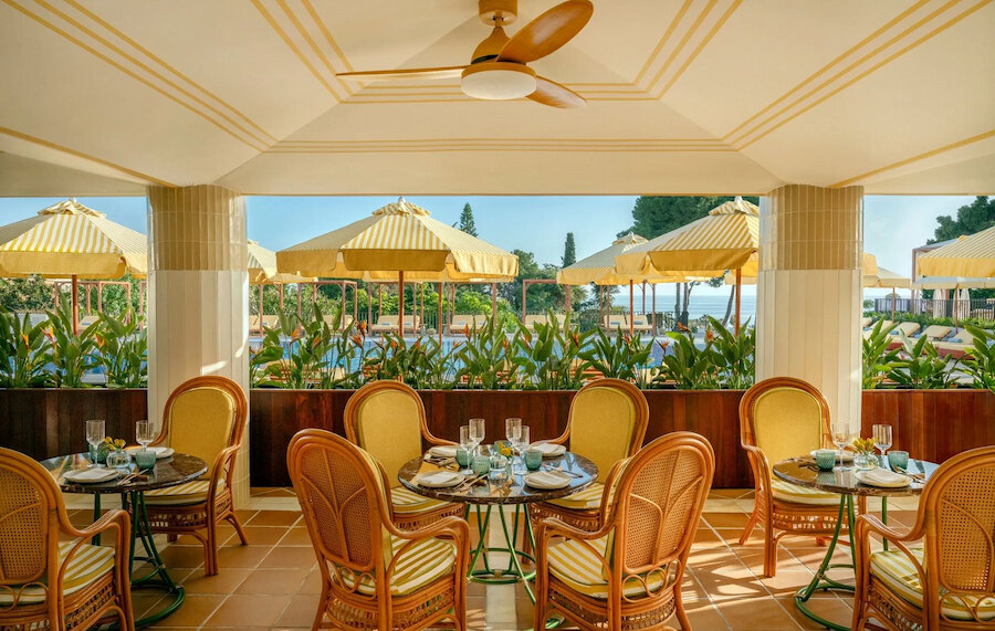 Licht en elegant restaurantterras met rotan stoelen, ronde marmeren tafels, geel gestreepte parasols en uitzicht op het zwembad in een verfijnde resortsetting.