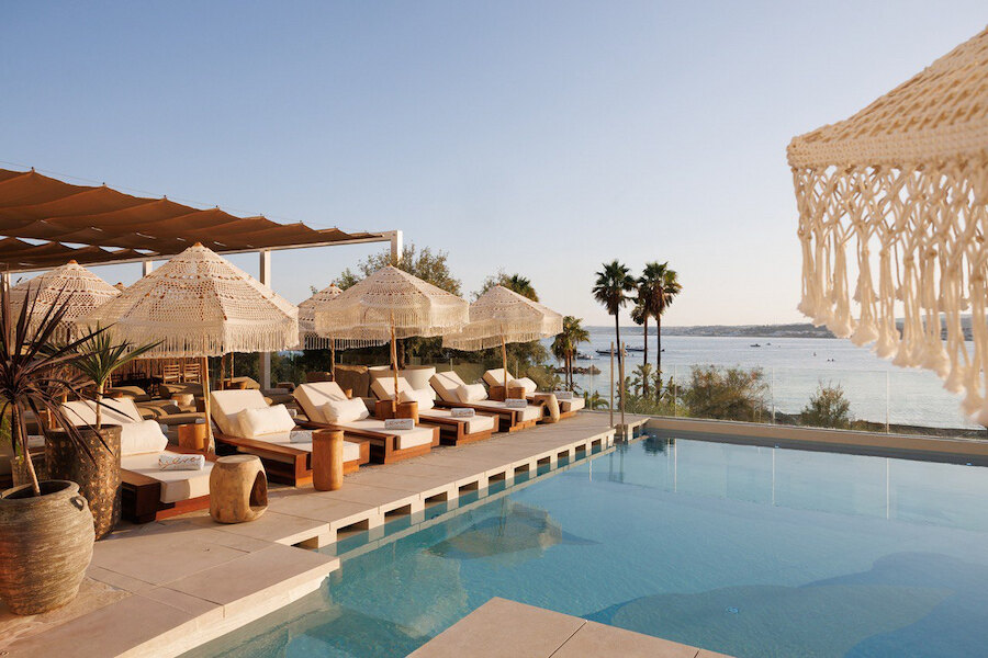 Stilvoller Poolbereich eines Beach Clubs mit gepolsterten Liegen, Sonnenschirmen mit Fransen, Palmen und ruhigem Meerblick in einem luxuriösen Ambiente direkt am Wasser.