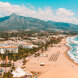 Vue aérienne du littoral de Marbella, avec une large plage de sable, la mer Méditerranée turquoise, une promenade bordée de palmiers, des bâtiments blancs en bord de mer, et la montagne de La Concha s’élevant à l’arrière-plan sous un ciel bleu lumineux.