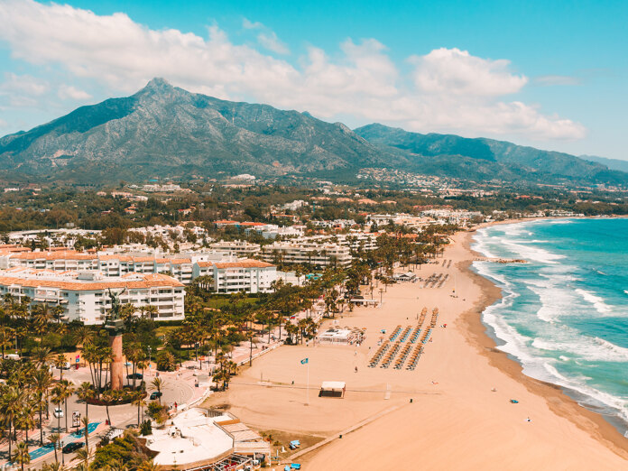 Aerial view of Marbella’s coastline with a wide sandy beach, turquoise Mediterranean Sea, palm-lined seafront, white beachfront buildings, and La Concha mountain rising in the background under a bright blue sky.