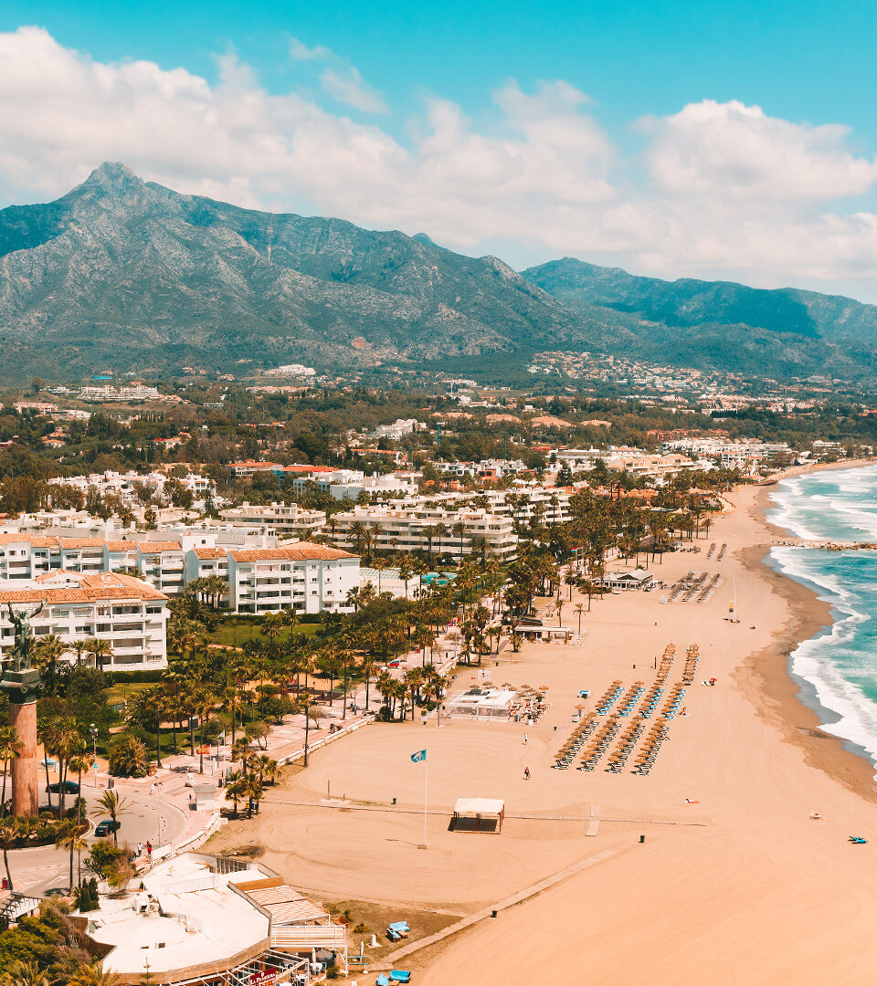 Luchtfoto van de kustlijn van Marbella met een breed zandstrand, de turquoise Middellandse Zee, een met palmen omzoomde boulevard, witte gebouwen aan het strand en de berg La Concha die op de achtergrond oprijst onder een helderblauwe lucht.
