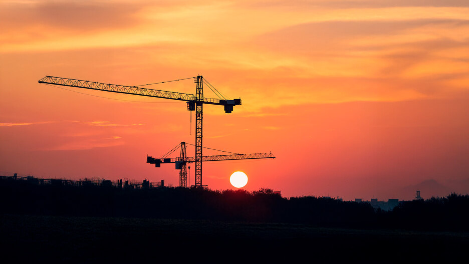Grúas de construcción recortadas sobre un intenso atardecer naranja, con el sol bajo en el horizonte sobre un atardecer.