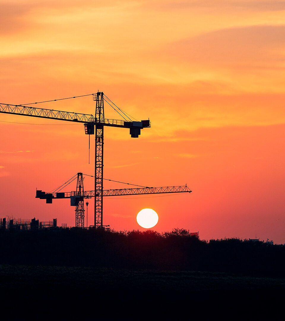 Construction cranes silhouetted against a vivid orange sunset, with the sun low on the horizon above a dark landscape.