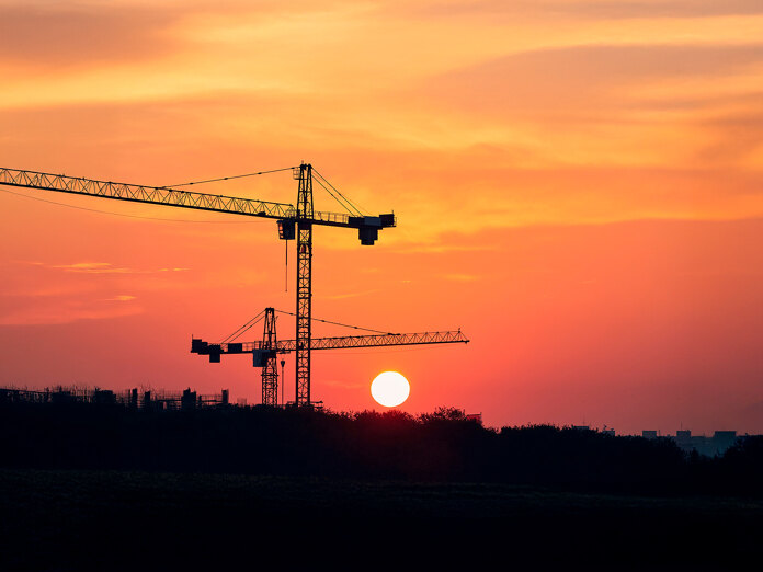 Bouwkranen als silhouetten tegen een feloranje zonsondergang, met de zon laag aan de horizon boven een donker landschap.