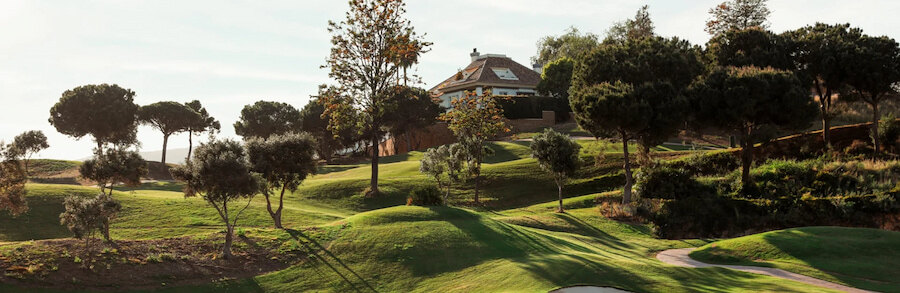 Los Naranjos Golf Club bij zonsondergang, met palmbomen, waterpartijen en uitzicht op de bergen.
