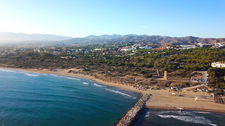 Luftaufnahme der Küste von Marbella mit Sandstränden, türkisfarbenem Wasser und den Bergen der Sierra Blanca im Hintergrund.