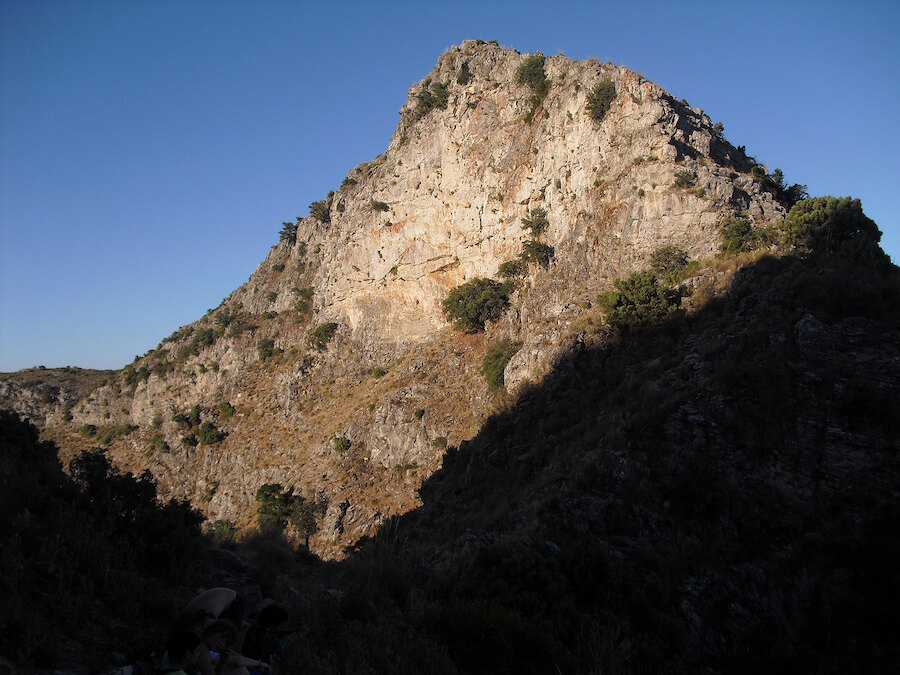 Door de zon verlichte kalkstenen top die boven een schaduwrijke vallei met verspreide mediterrane begroeiing uitrijst.