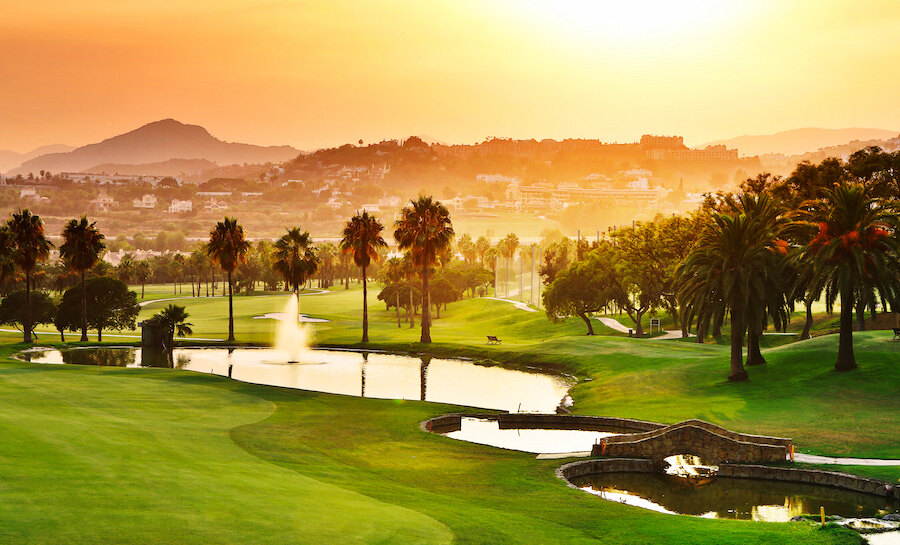 Los Naranjos Golf Club bij zonsondergang, met palmbomen, waterpartijen en uitzicht op de bergen.