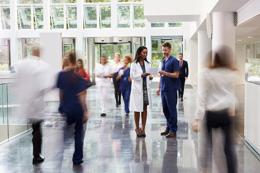 Medical professionals talking in a bright hospital lobby, with staff and visitors moving through the space.
