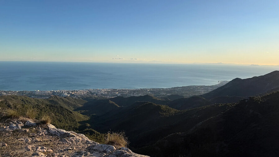 Panoramic view of Marbella’s coastline and the Mediterranean Sea from a mountain summit, with rolling hills in the foreground under a clear sky.