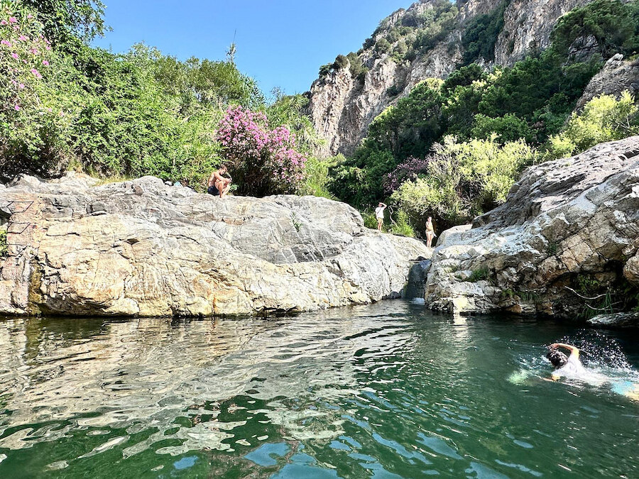 Natural rock pools surrounded by steep cliffs and lush greenery, with people swimming and relaxing in a peaceful mountain setting near Marbella.