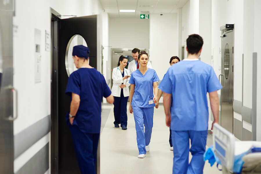 Hospital staff walking through a clean clinical corridor, including nurses and doctors in scrubs.