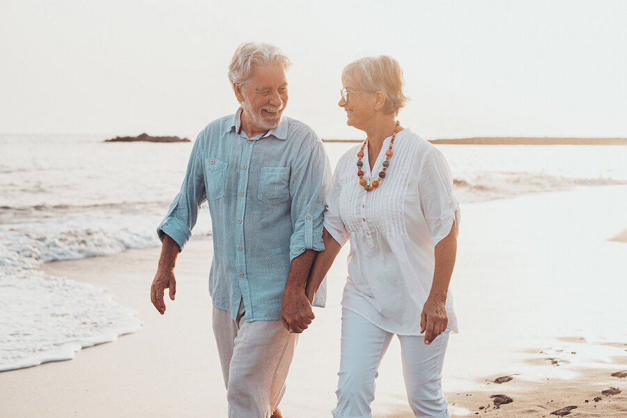 Una pareja mayor feliz caminando de la mano por una playa soleada, sonriéndose mutuamente con suaves olas al fondo.