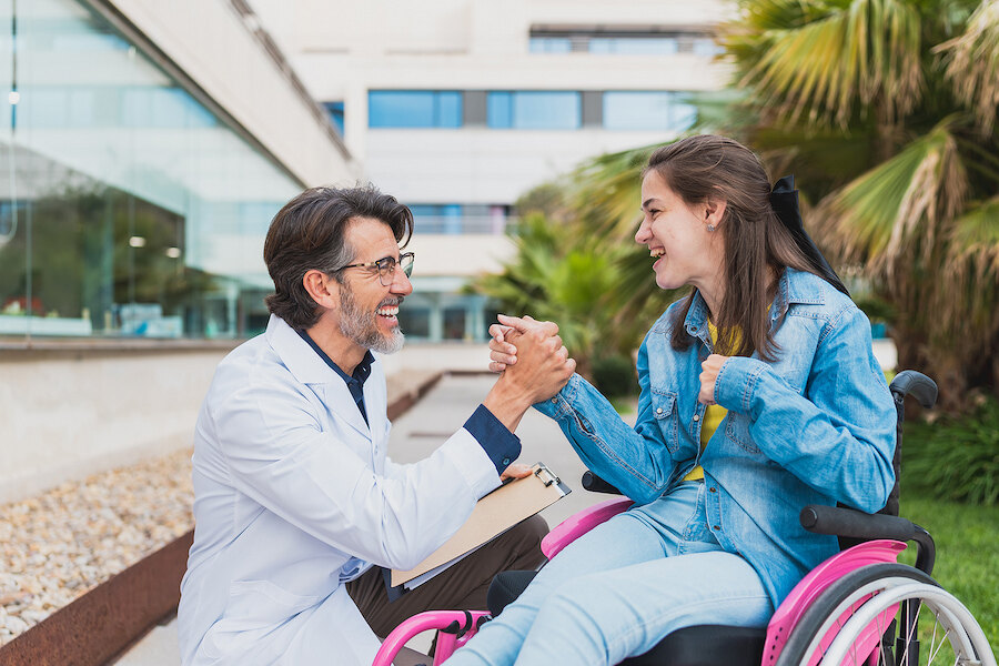 Doctor greeting a smiling young woman in a wheelchair outside a modern medical facility.
