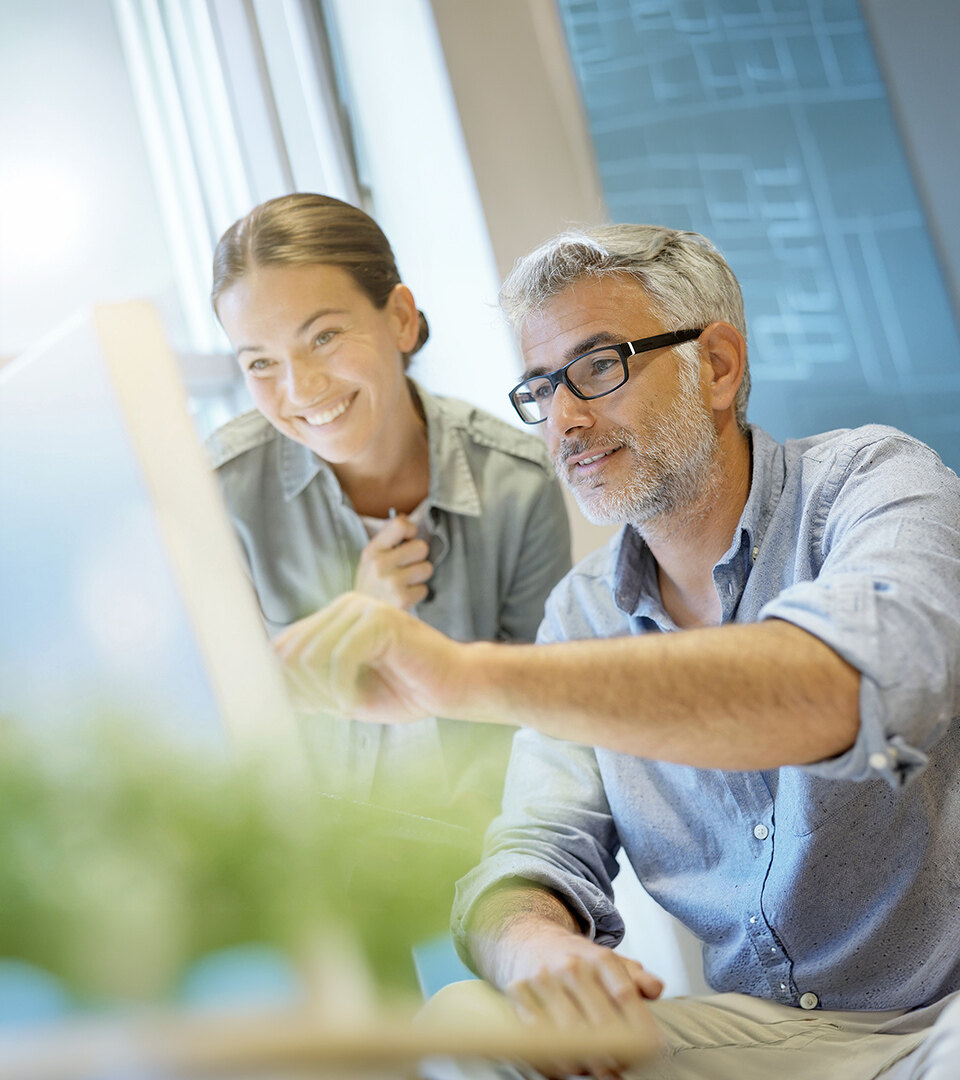 Dos compañeros colaborando frente a un ordenador en una oficina moderna, con un hombre señalando la pantalla mientras una mujer sonríe a su lado, rodeados de luz natural y vegetación.