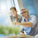 Best time to buy. Two colleagues collaborating at a computer in a modern office, with a man pointing at the screen while a woman smiles beside him, surrounded by natural light and greenery.