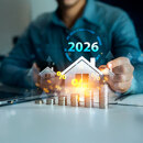2026 real estate market concept with house icon, rising coin stacks, percentage symbols and a person working at a desk with a laptop.