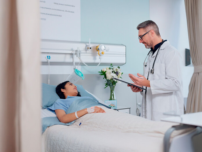 Een arts spreekt met een patiënt die in een ziekenhuisbed ligt, in een lichte privékamer.