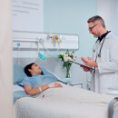 Doctor speaking with a patient lying in a hospital bed in a bright private room.