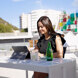 Una mujer trabajando en una tablet en una terraza exterior soleada, con arquitectura moderna y un edificio colorido al fondo.
