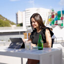 A woman working on a tablet at a sunny outdoor terrace, with modern architecture and a colourful building in the background.