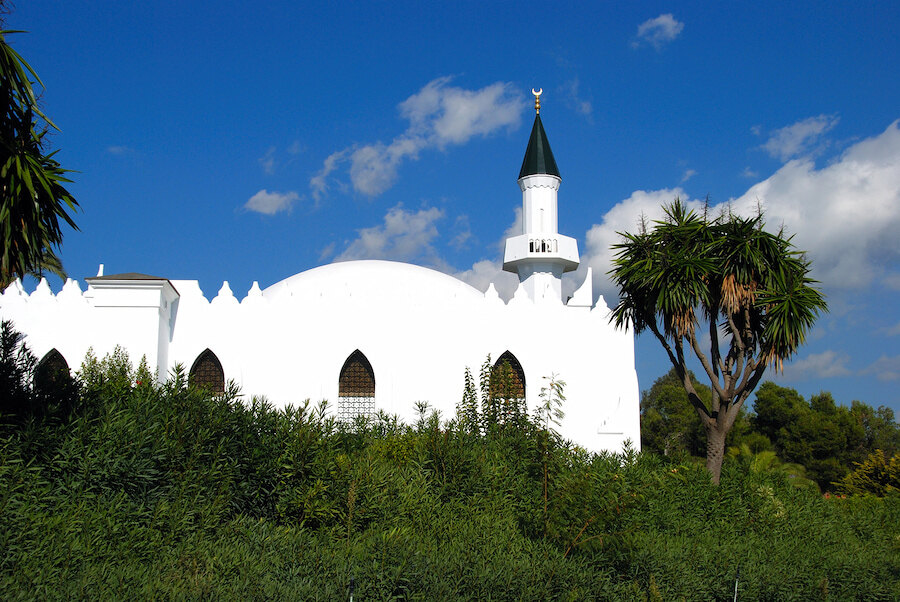 White mosque with a green-topped minaret and crescent symbol, surrounded by lush greenery and palm trees under a clear blue sky.