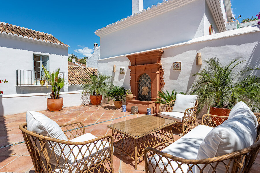 Terrasse ensoleillée sur le toit d’une maison mitoyenne du Golden Mile de Marbella, avec sols en terre cuite, mobilier de salon en osier, palmiers en pots et fontaine murale décorative contre des murs blanchis à la chaux.