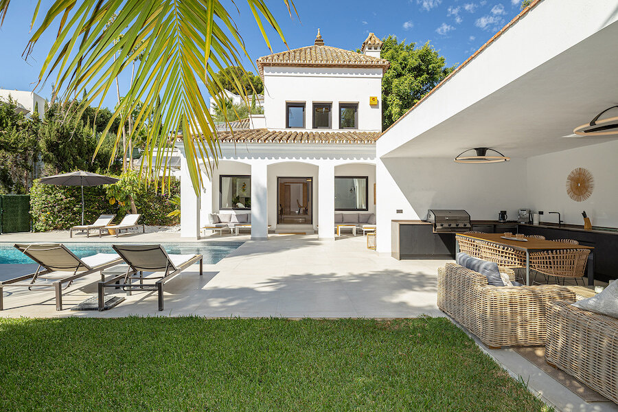 Sunny villa terrace with swimming pool, sun loungers and covered outdoor dining area with built-in barbecue, set beside a white Mediterranean-style home.