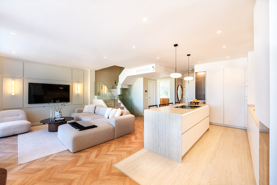 Espace de vie moderne à aire ouverte dans une maison mitoyenne du Golden Mile de Marbella, avec îlot de cuisine minimaliste, parquet en chevrons, canapés aux tons neutres et escalier en verre.
