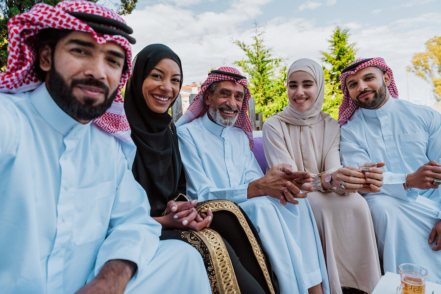 Group of five people wearing traditional Middle Eastern attire sitting outdoors together, smiling and holding small cups, enjoying a social gathering in a garden setting.