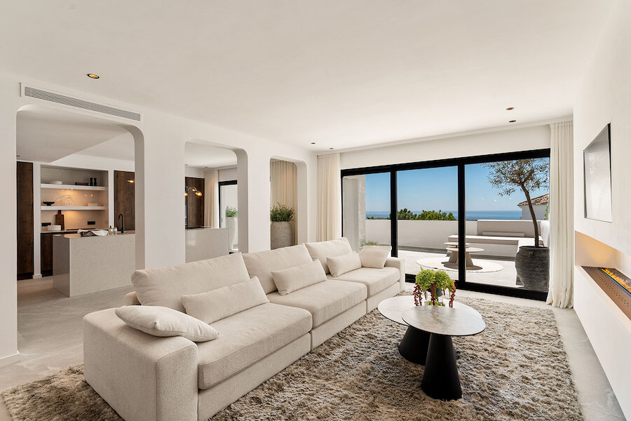 Bright open-plan living room with neutral sofas and modern fireplace, opening onto a terrace with sea views through large sliding glass doors.