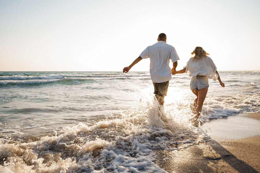 Couple se tenant la main et courant le long du rivage au coucher du soleil, éclaboussant les vagues sur une plage de sable.