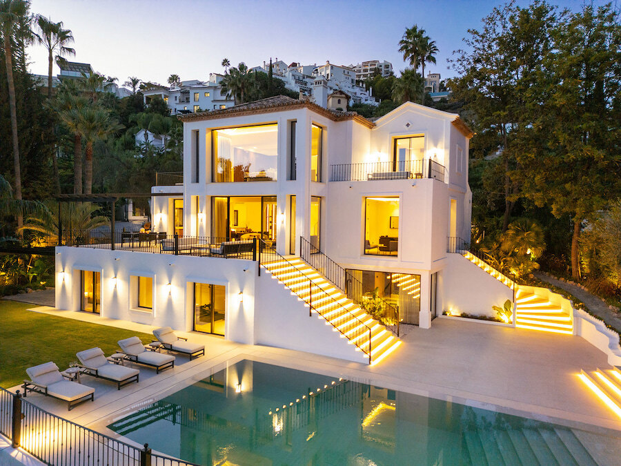 Modern white villa at dusk with illuminated staircases and large windows, overlooking a swimming pool and sun terrace in a landscaped garden.