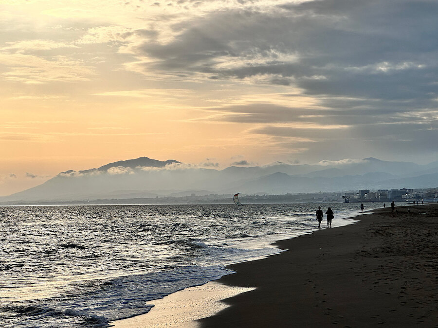 Moody sunset view along Marbella’s shoreline, with gentle waves, silhouettes of people walking on the beach, and misty mountains in the distance.