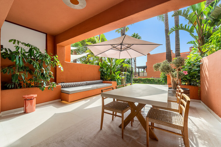 Terrasse couverte aux murs en terre cuite sur le Golden Mile de Marbella, avec banquette intégrée, table à manger entourée de chaises tressées, grand parasol et jardin tropical luxuriant en arrière-plan.