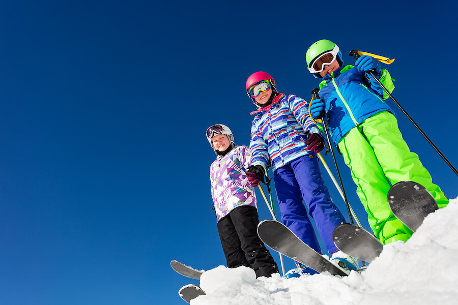Trois enfants en tenues de ski colorées se tenant sur une piste enneigée avec skis et bâtons, sous un ciel bleu clair.
